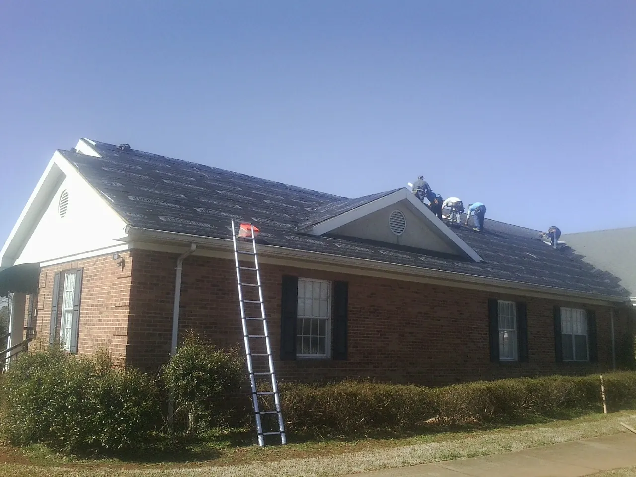 Crew tearing shingles off a brick ranch roof with a ladder set — Emergency Roof Repair work by our crew in Cameron